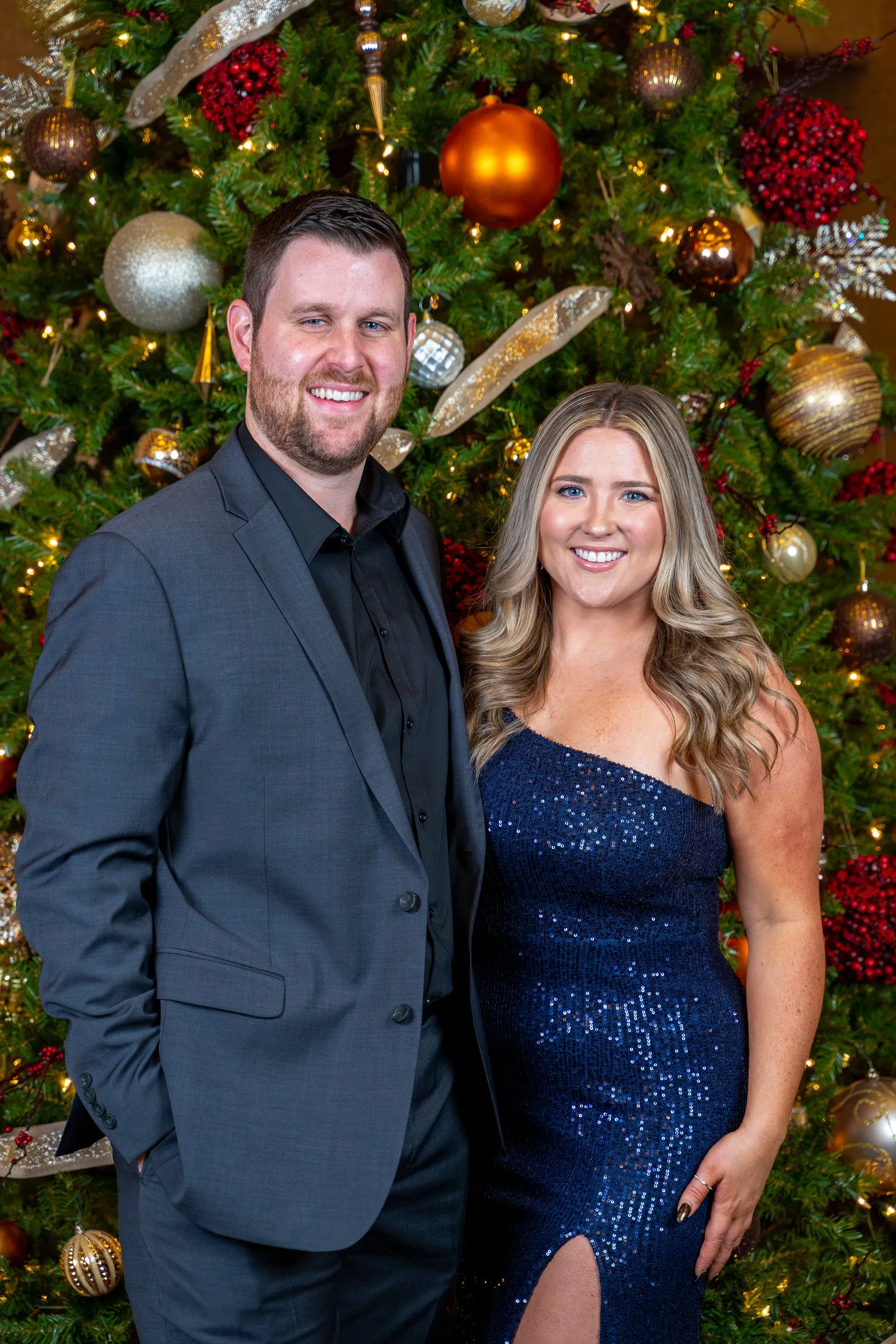 Man in a suit and woman in a navy sequin gown pose by a Christmas tree.