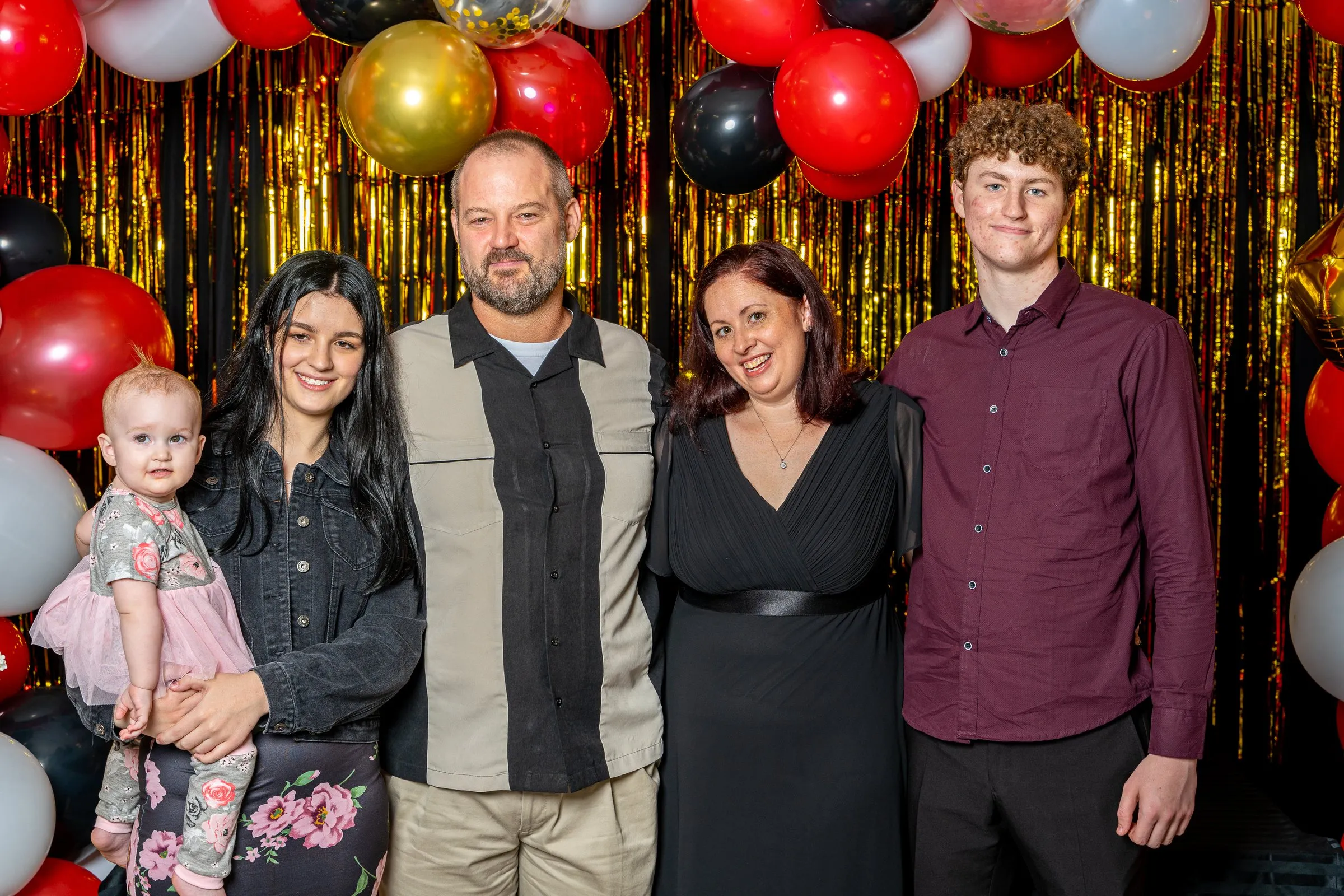 Five people, including a toddler, pose in front of a red, black and gold balloon garland.