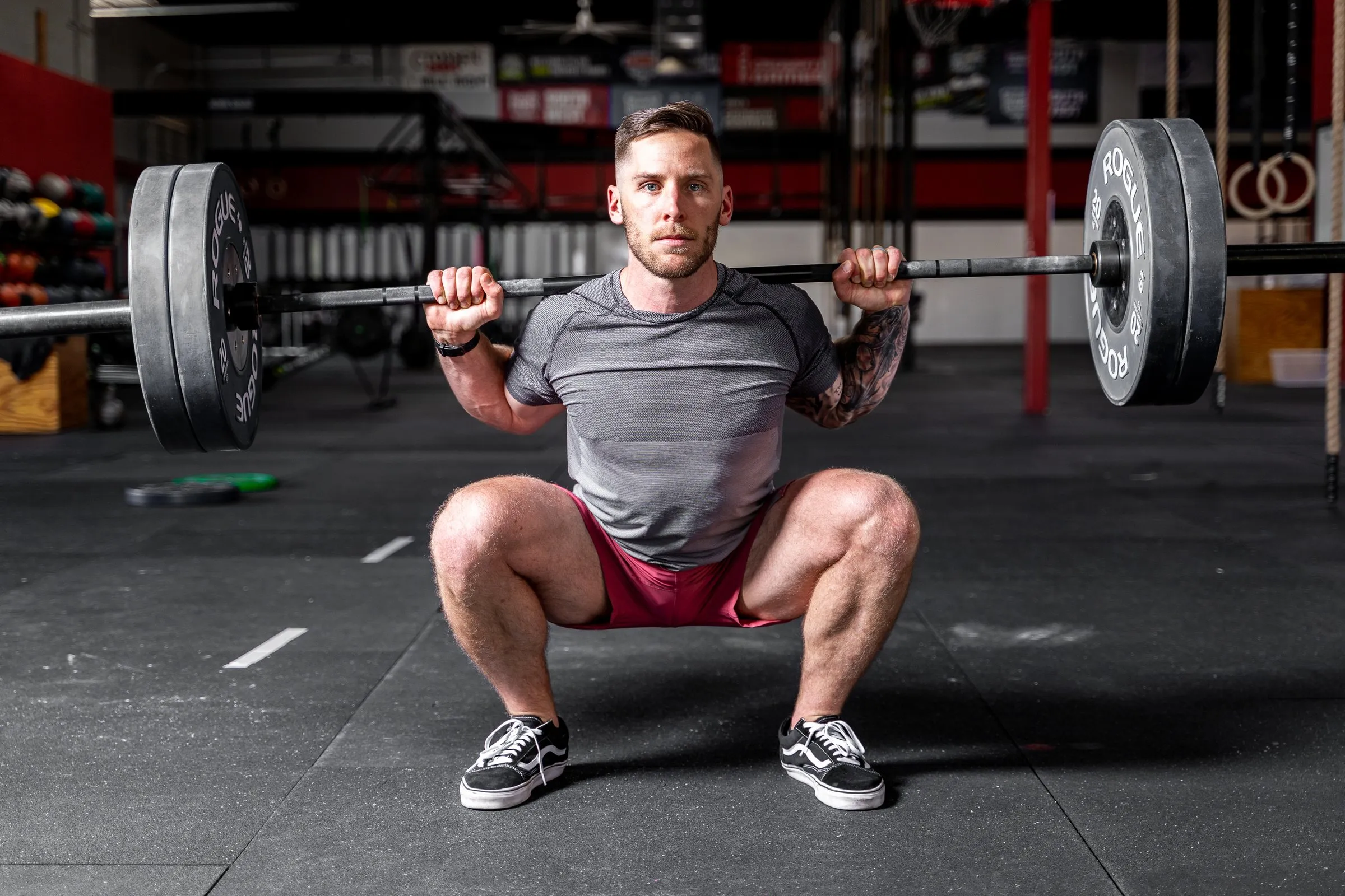 Man in a grey t-shirt performing a barbell back squat in a gym, facing the camera.