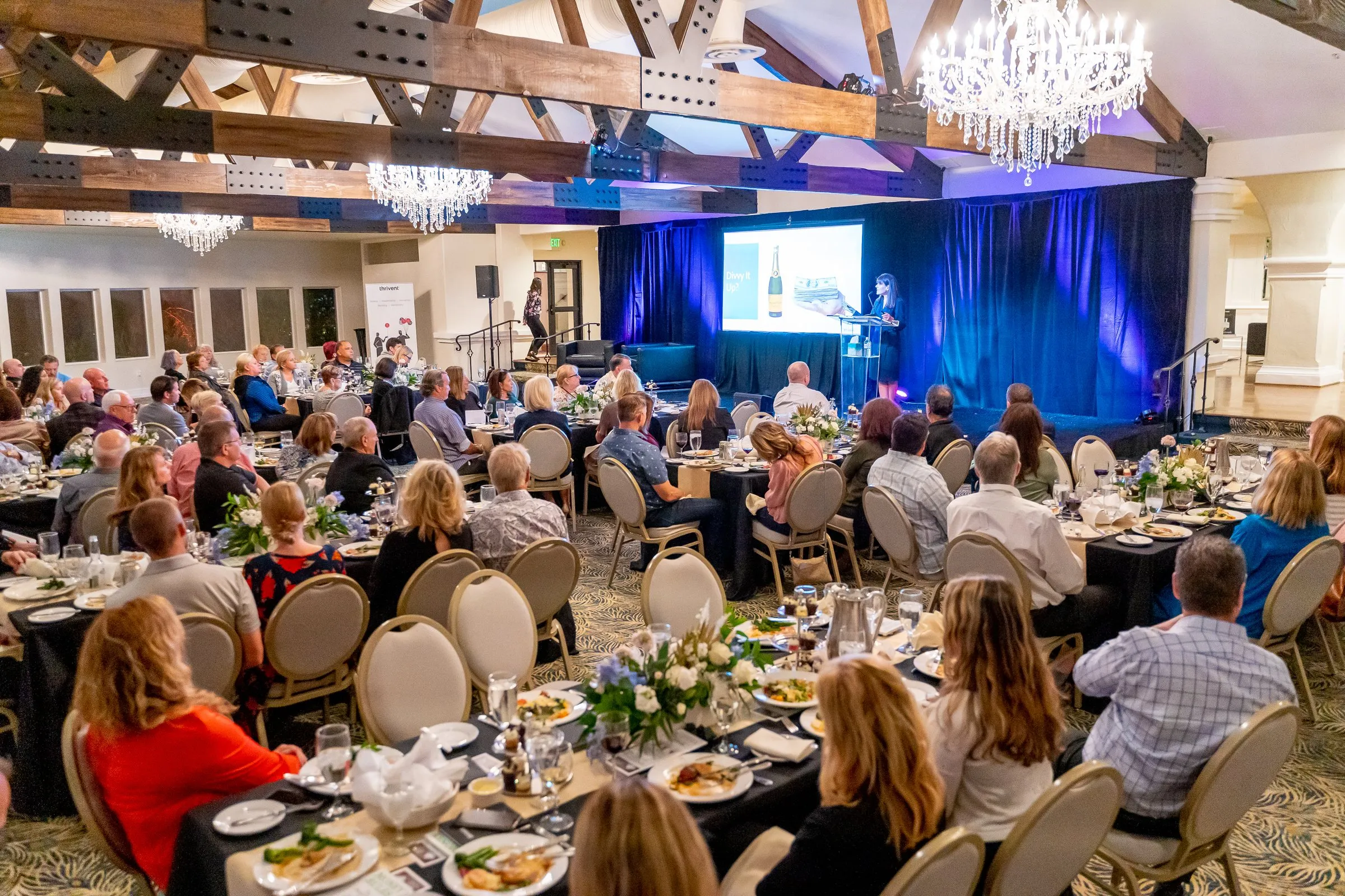Ballroom with chandelier, round tables of guests and a speaker on stage.