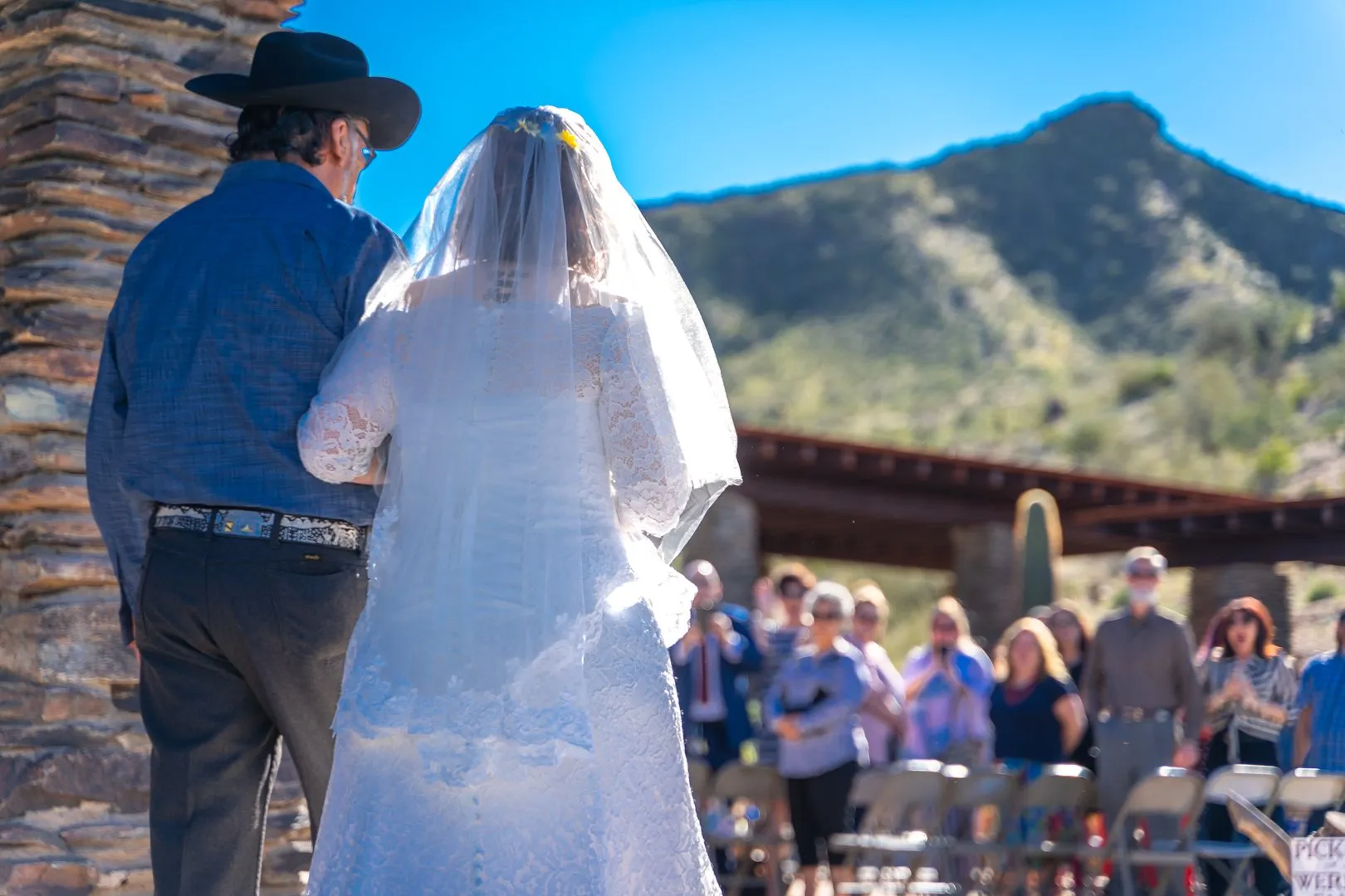 Veiled woman in a white gown walks beside a man toward guests at an outdoor ceremony.
