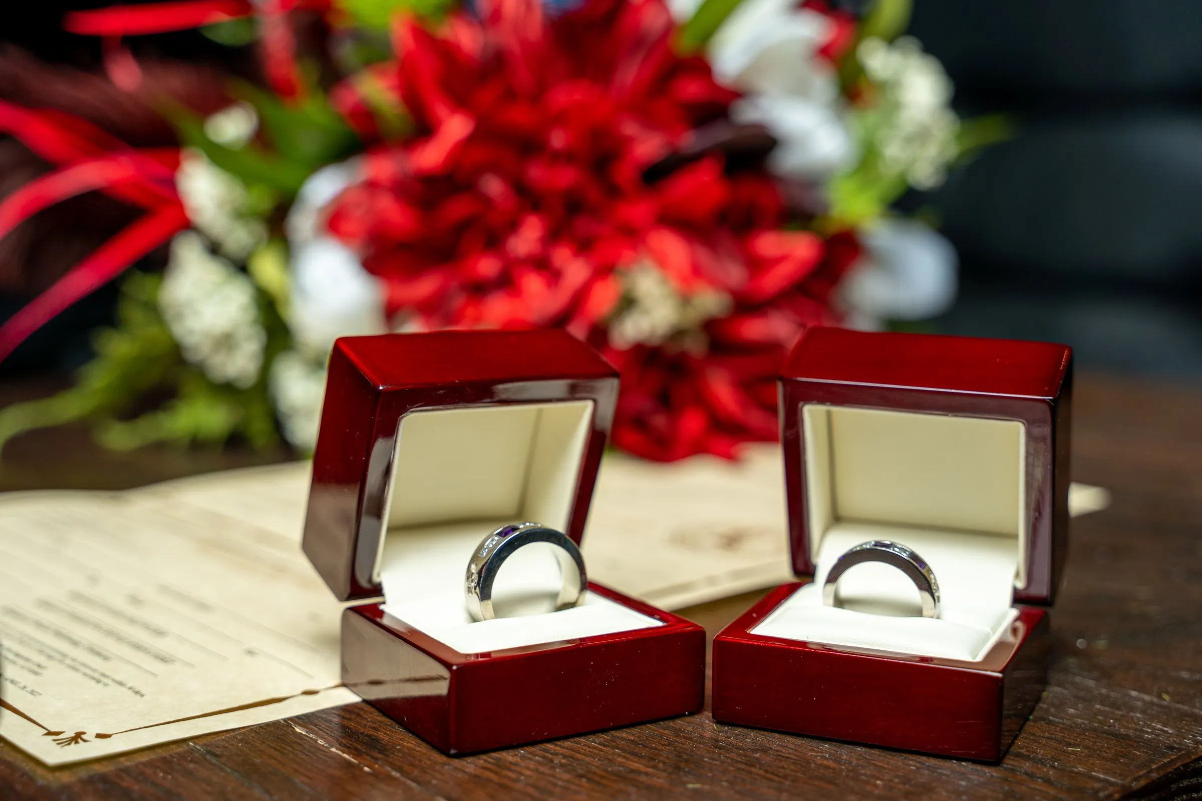 Two silver wedding rings in open red wooden boxes before a red floral bouquet.
