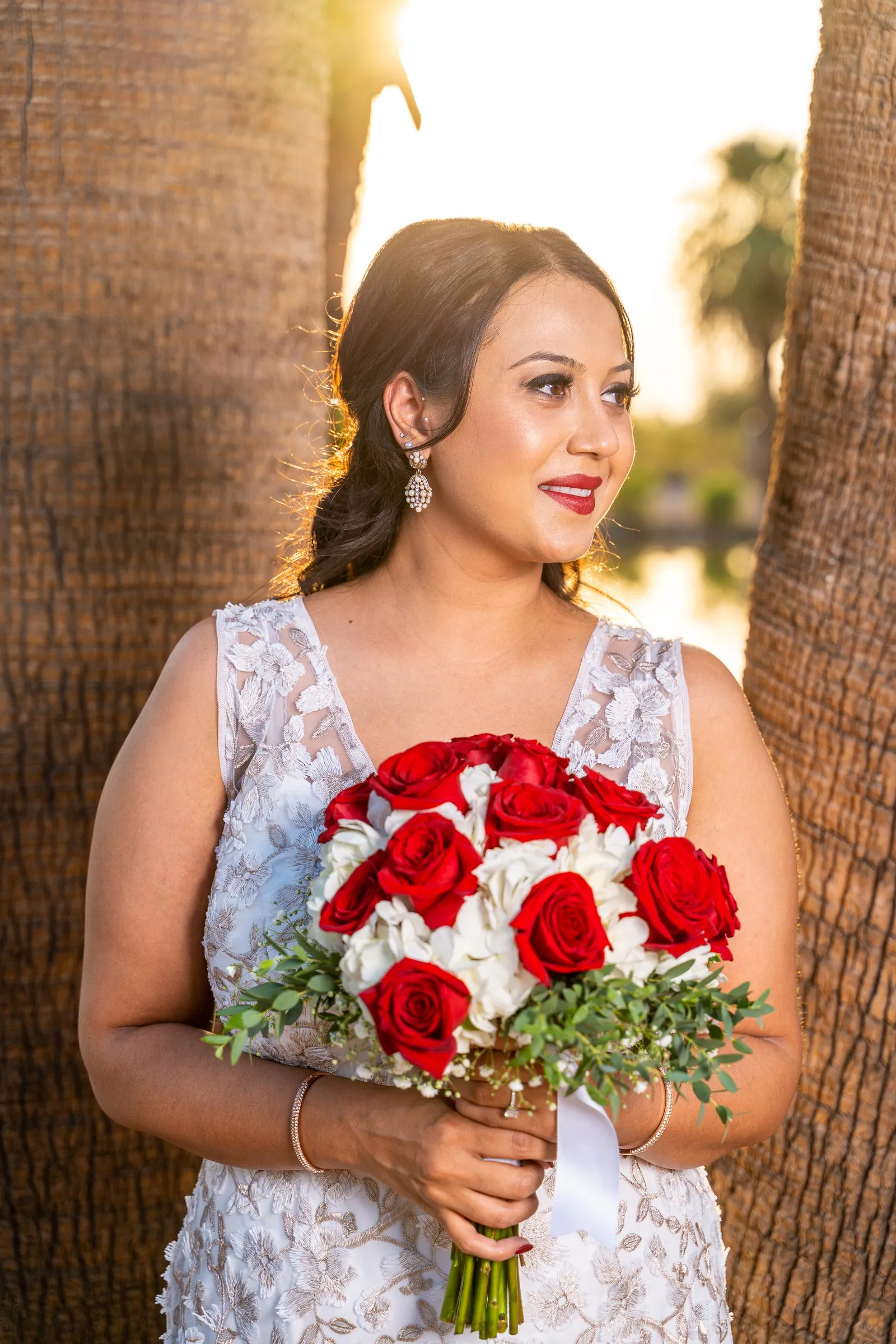 Woman in a white lace dress holds red roses between palm trunks at sunset.