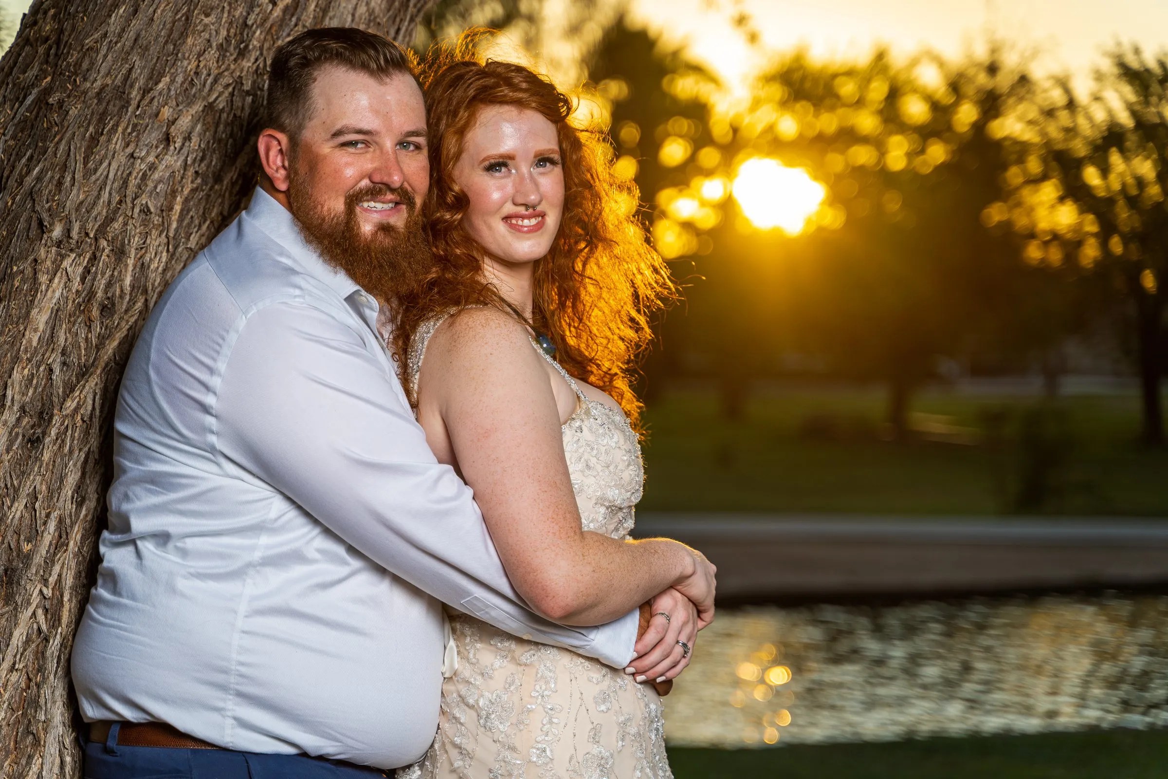 Man and woman in wedding attire embrace against a tree as the sun sets behind them.