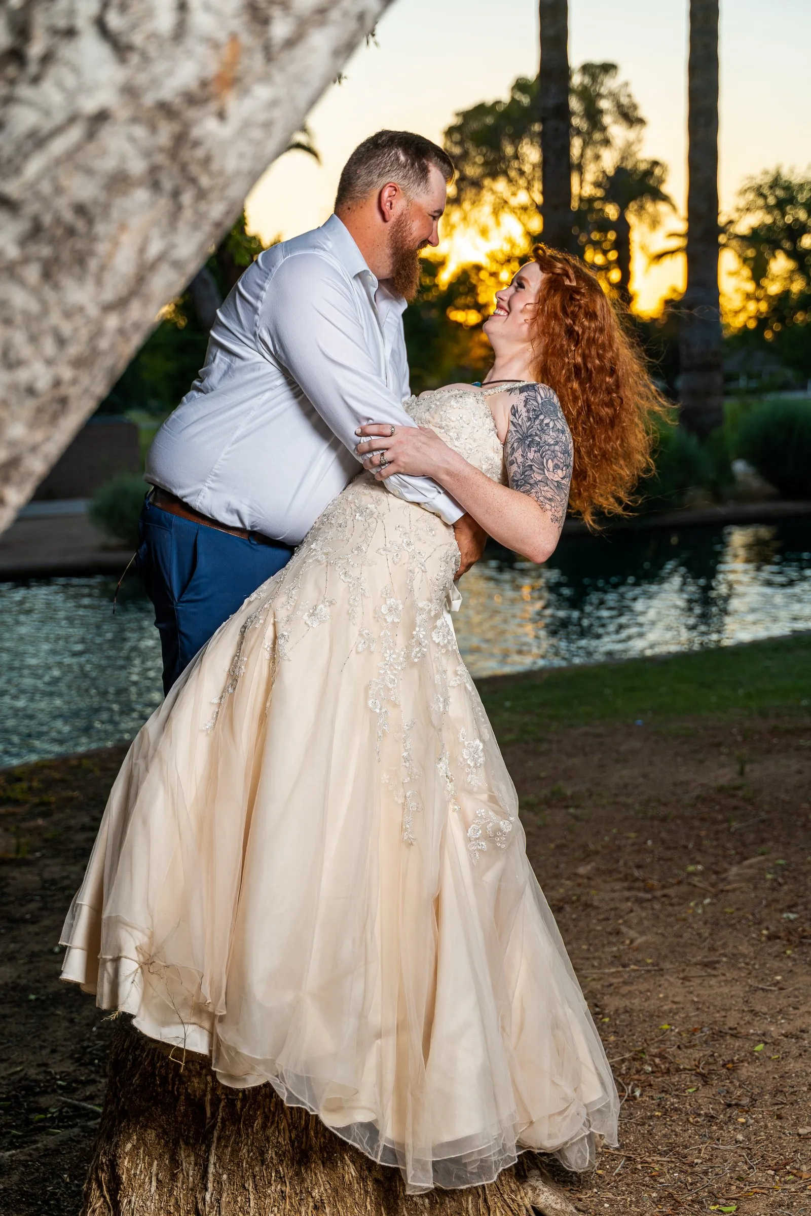 Man dips a woman in a cream gown backward beside a pond at sunset.