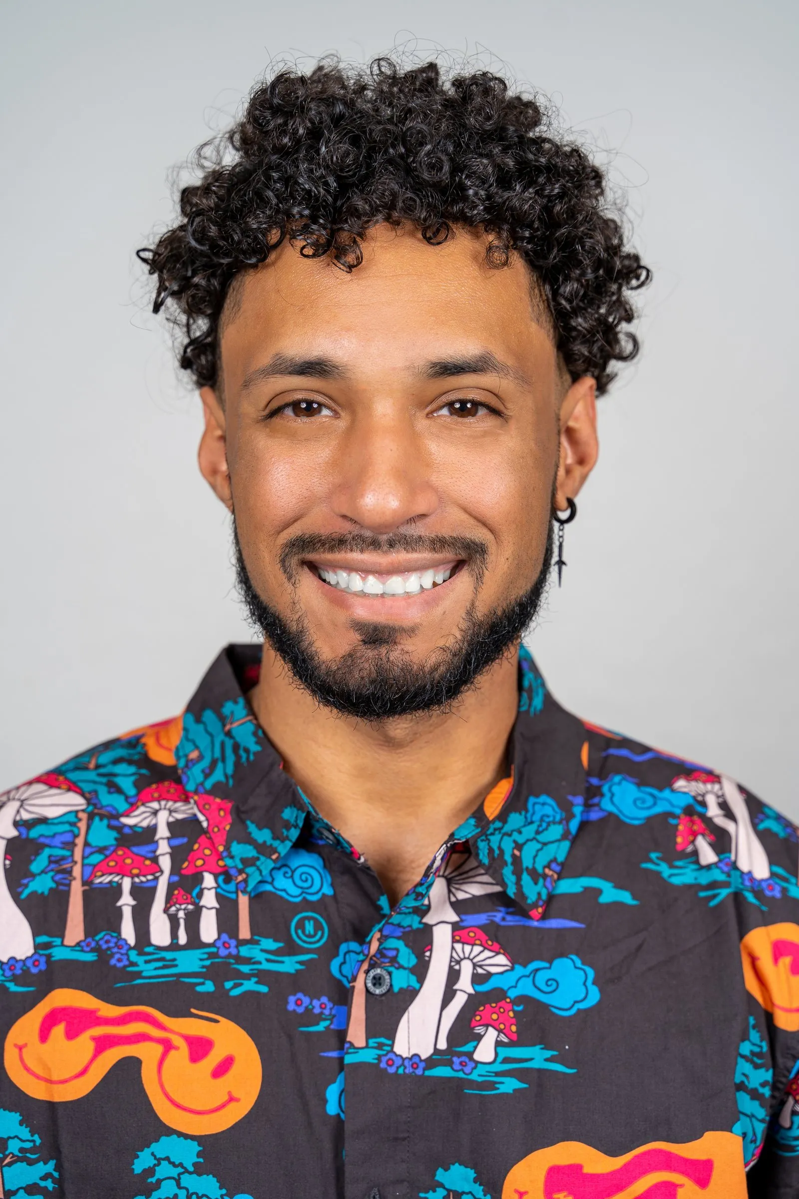 Smiling bearded man with curly hair in a mushroom-print shirt against a light gray backdrop.
