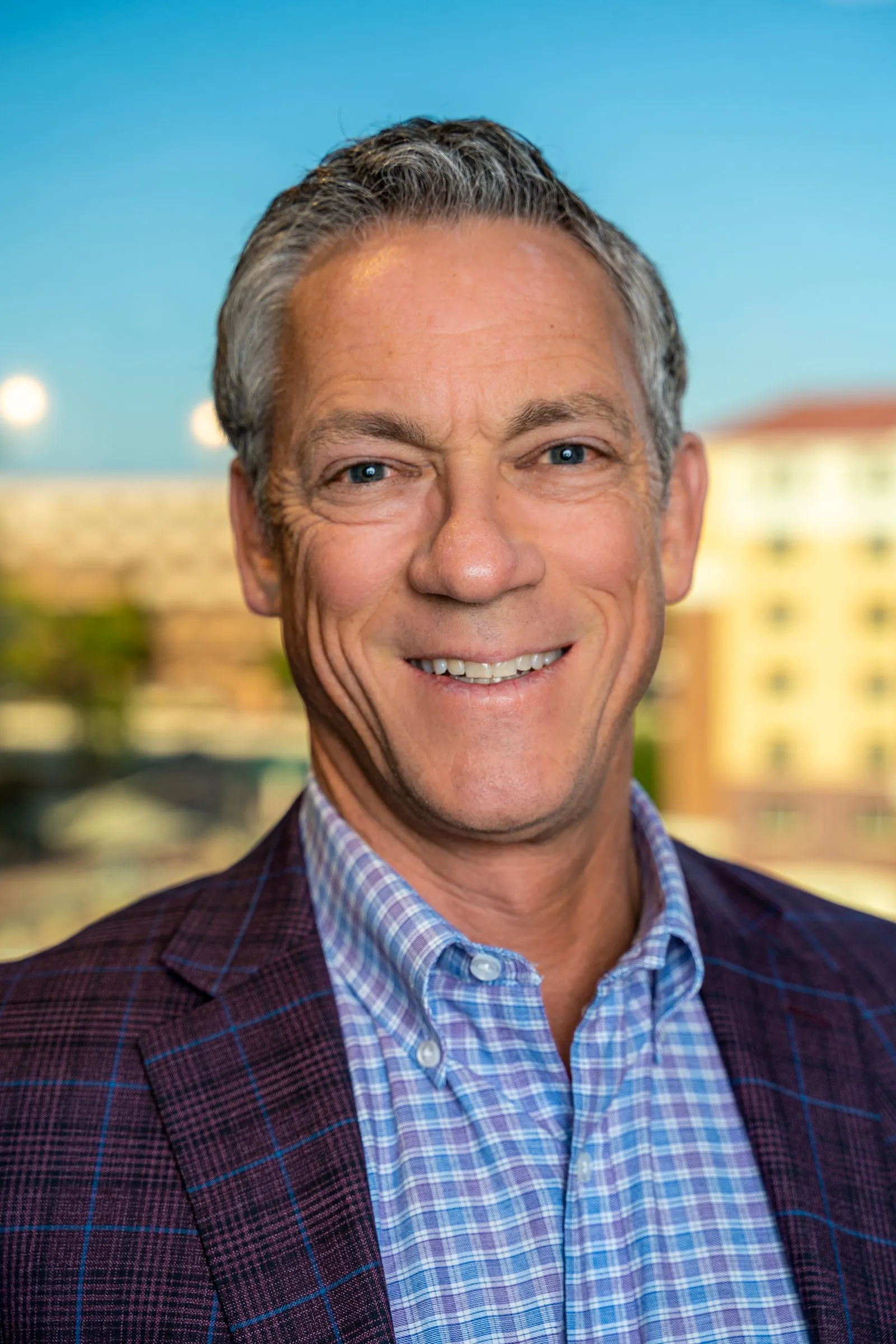 Smiling man with silver hair in a plaid blazer against a blurred sunlit skyline.