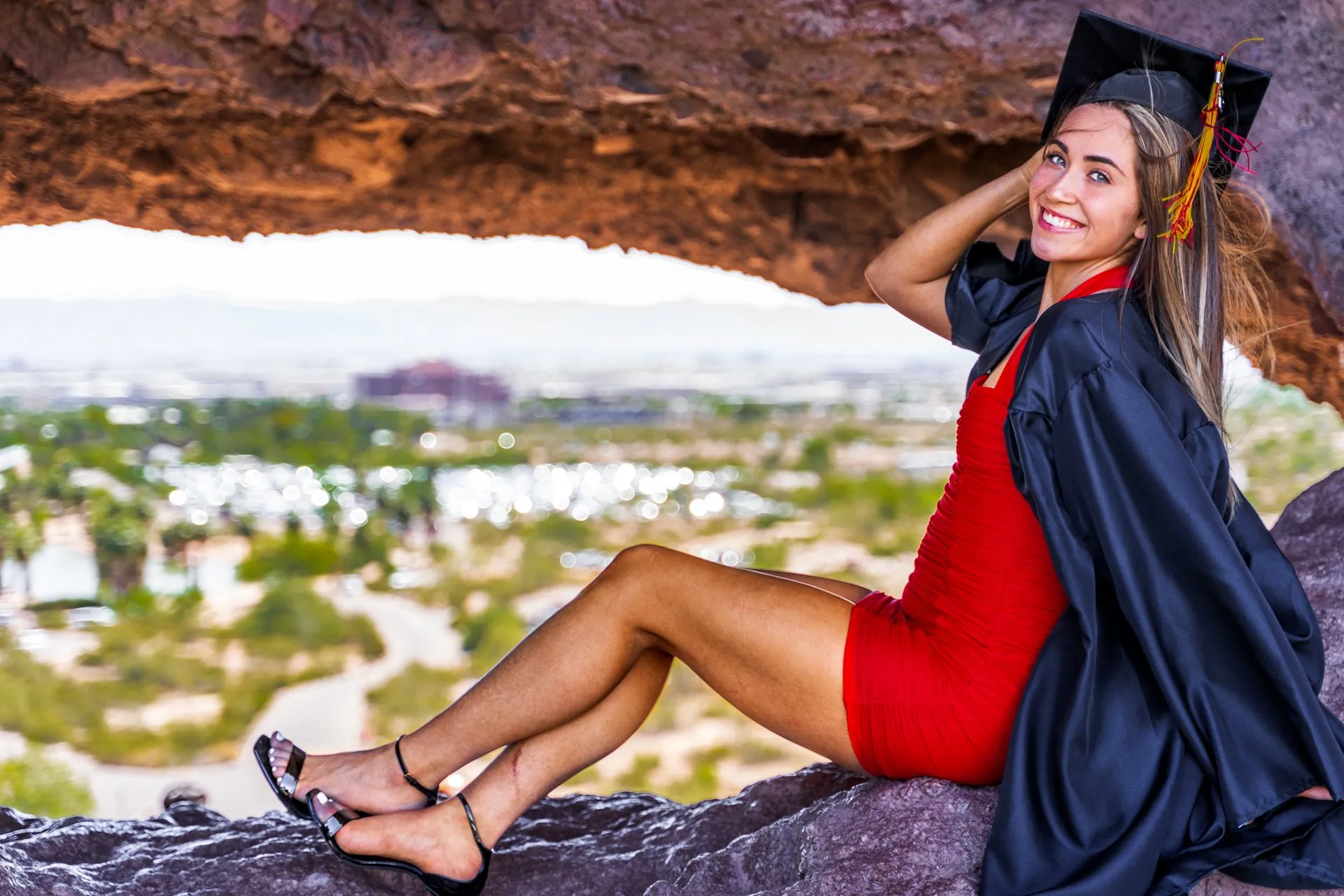 Graduate in cap and gown sits in a rocky opening with a city view behind.