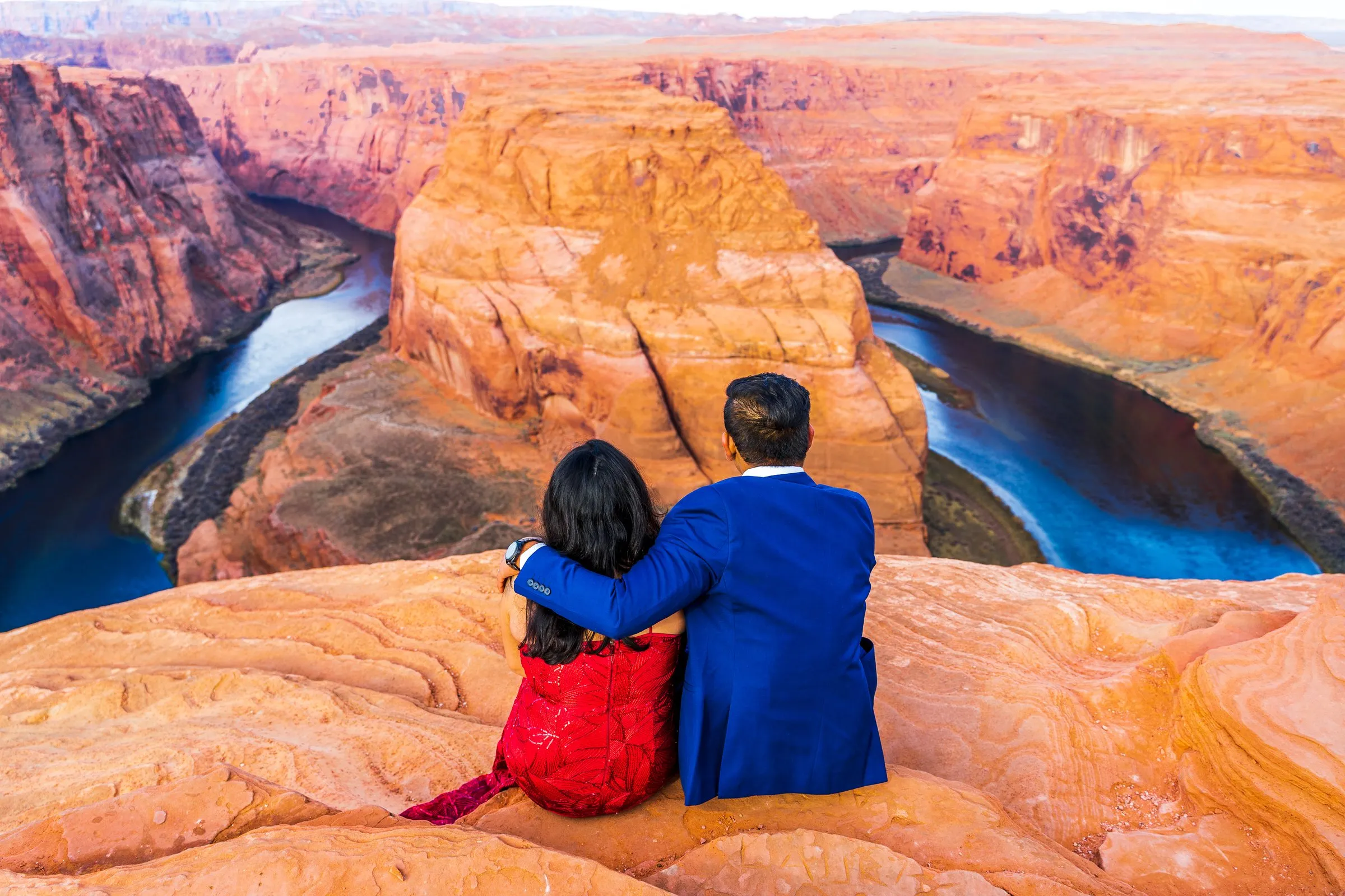 Two people sit close on an orange sandstone ledge overlooking a deep canyon and river bend.