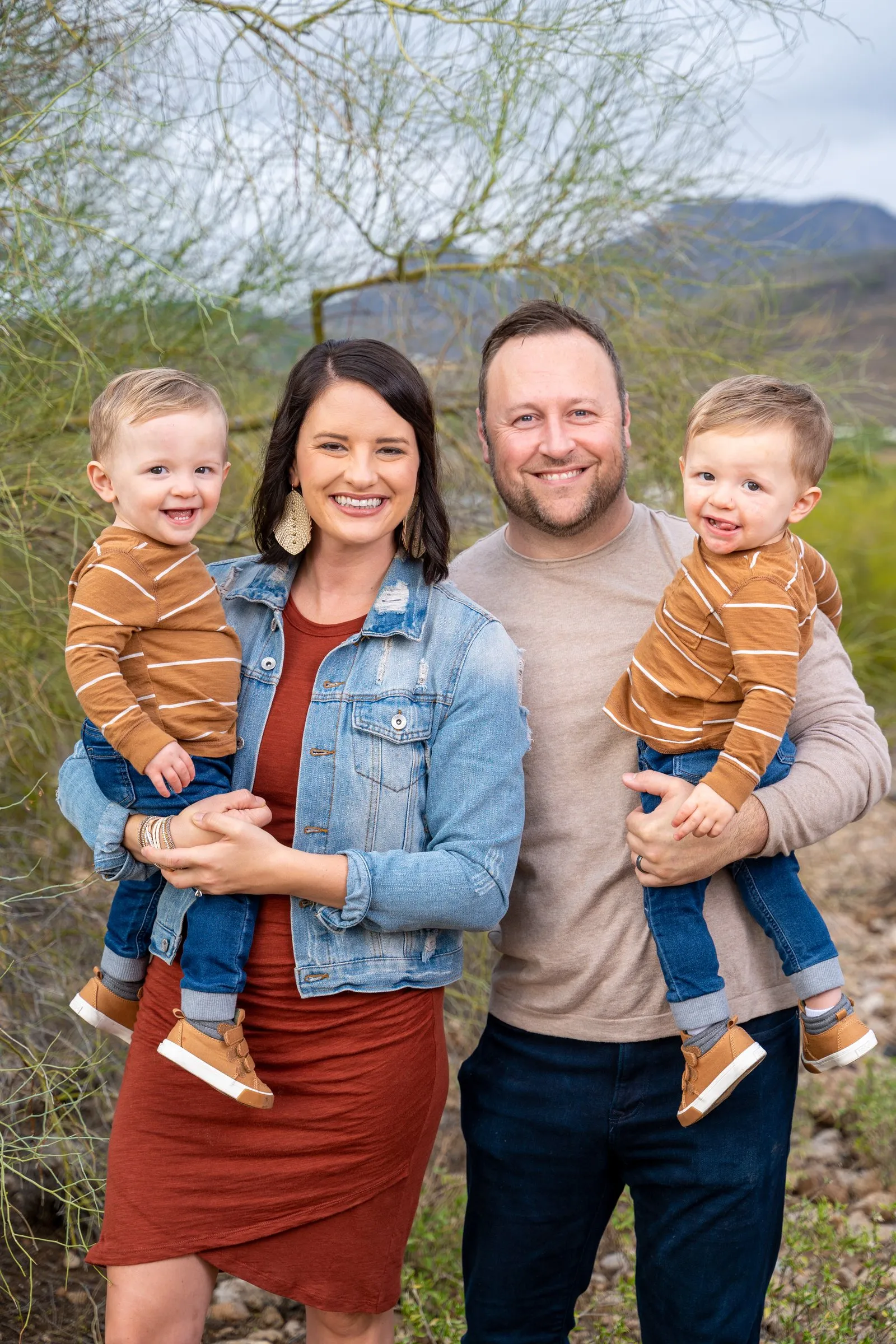 Smiling man and woman each hold a laughing toddler outdoors with hills behind.