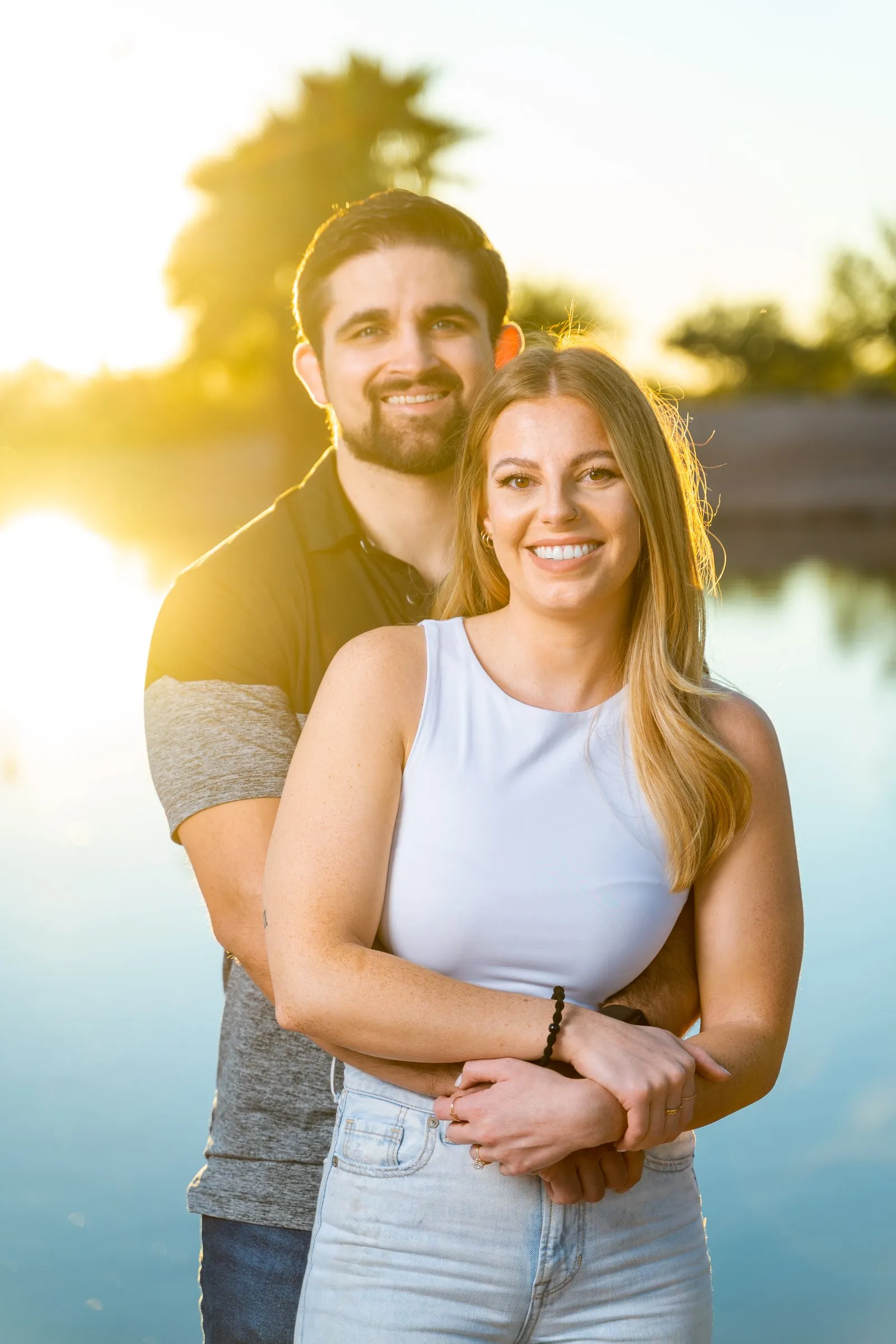 Smiling couple embrace in front of a lake lit by warm golden sunset light.