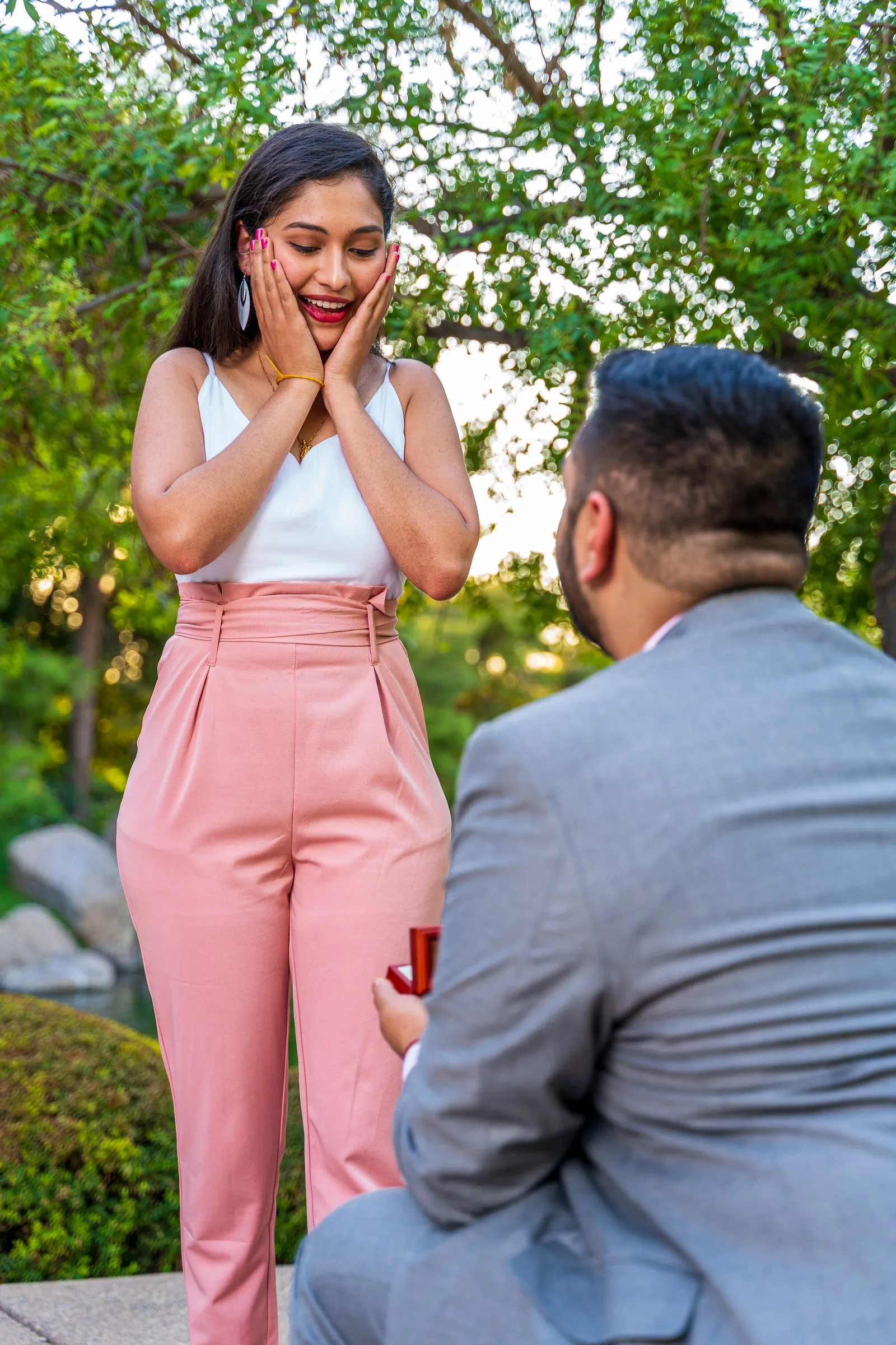 A woman clasps her cheeks in joyful surprise as a man kneels with a ring box.