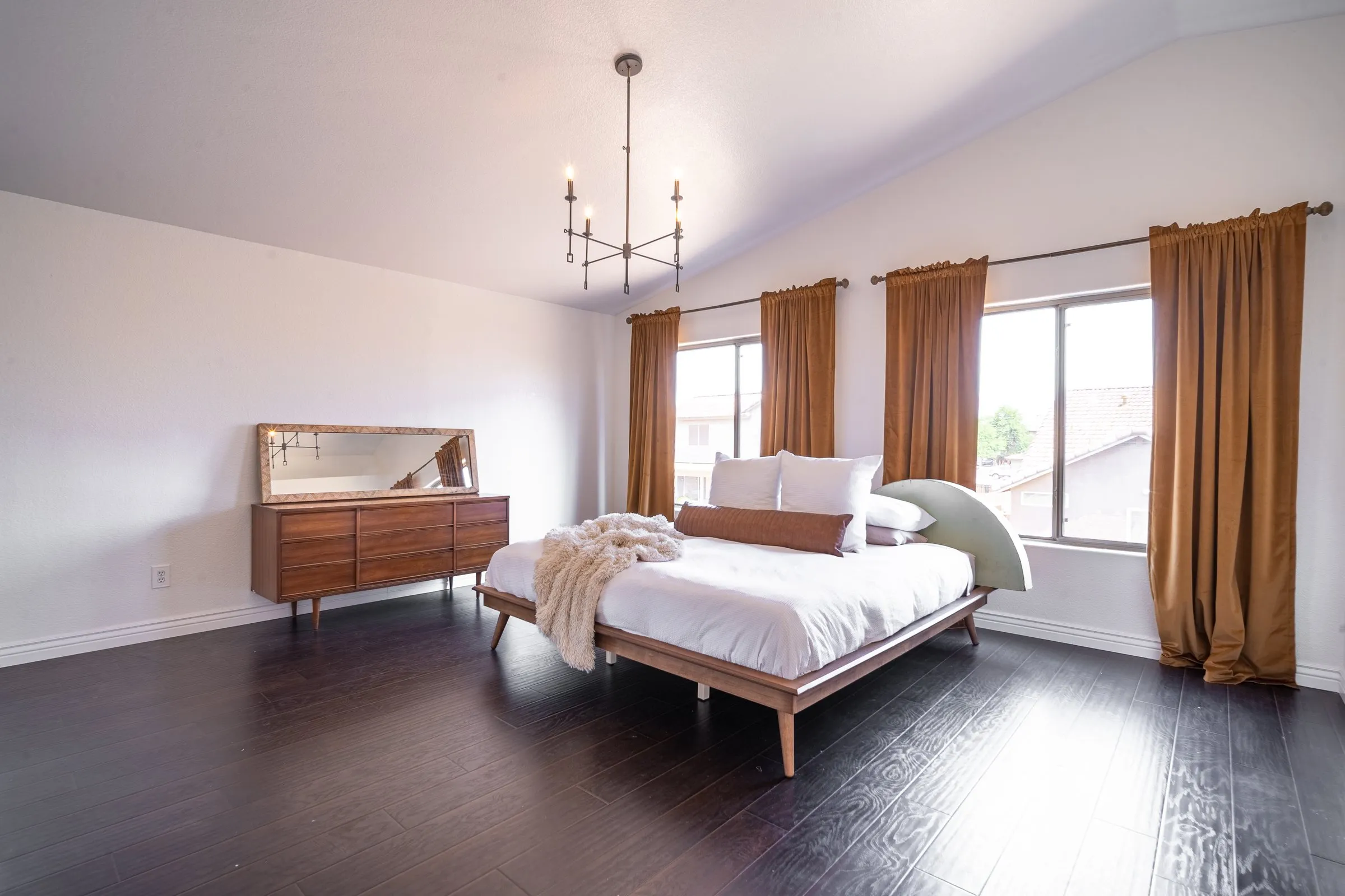Minimal bedroom with a wood platform bed, chandelier, copper curtains and dark floor.